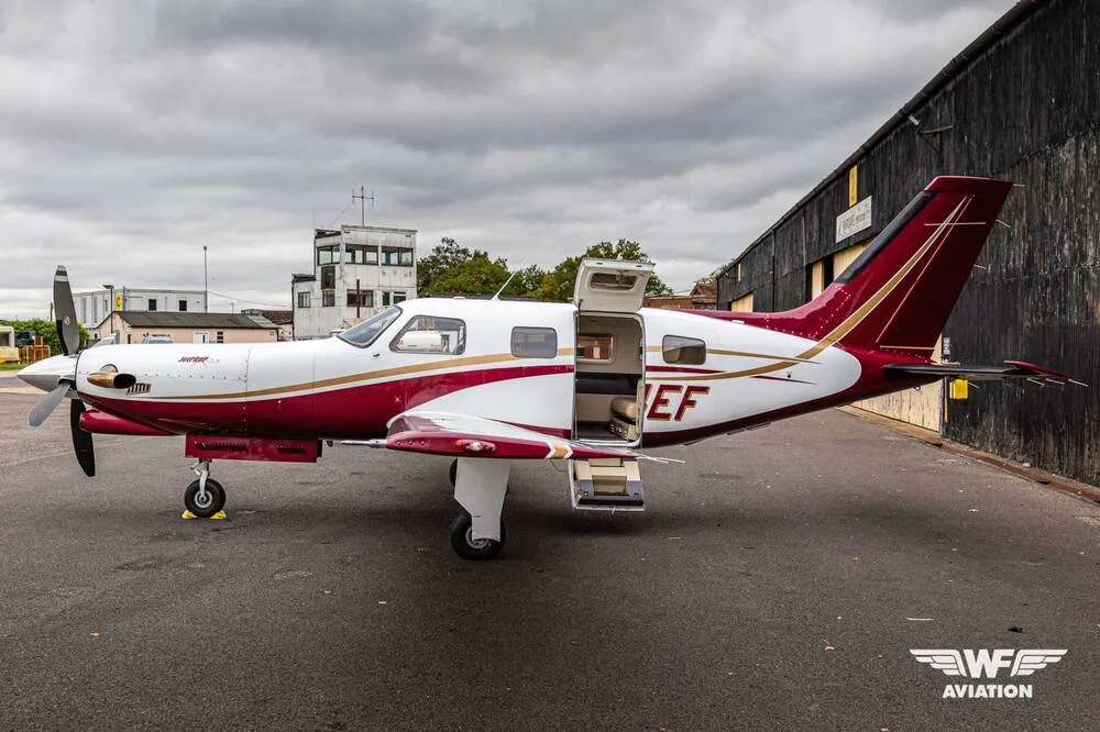 A side view of the N14EF turboprop aircraft with its main cabin door open, parked on a tarmac with an old control tower in the background.