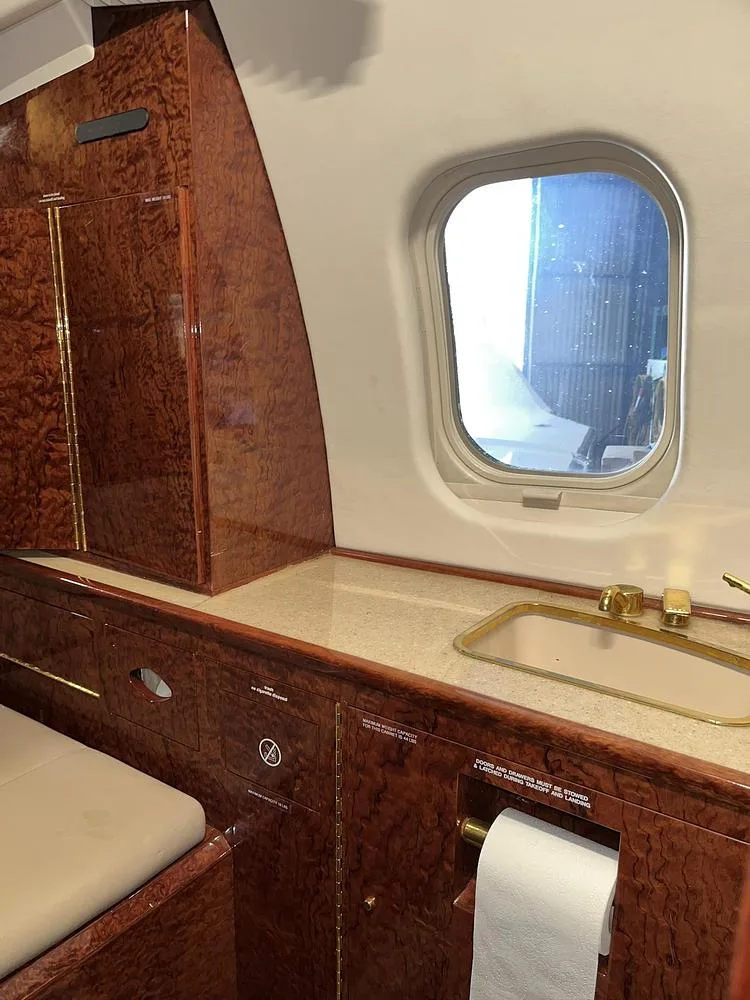 A private jet lavatory or galley area with a small sink, wood paneling, and a window.