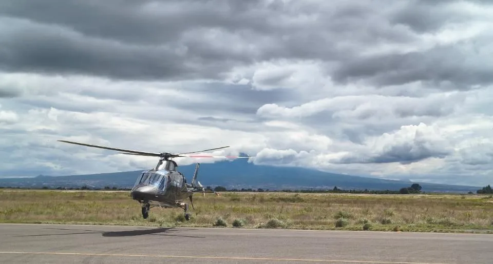 A dark grey Leonardo AW109 GrandNew helicopter hovers just above a runway in a grassy field with a large mountain in the background under a cloudy sky.