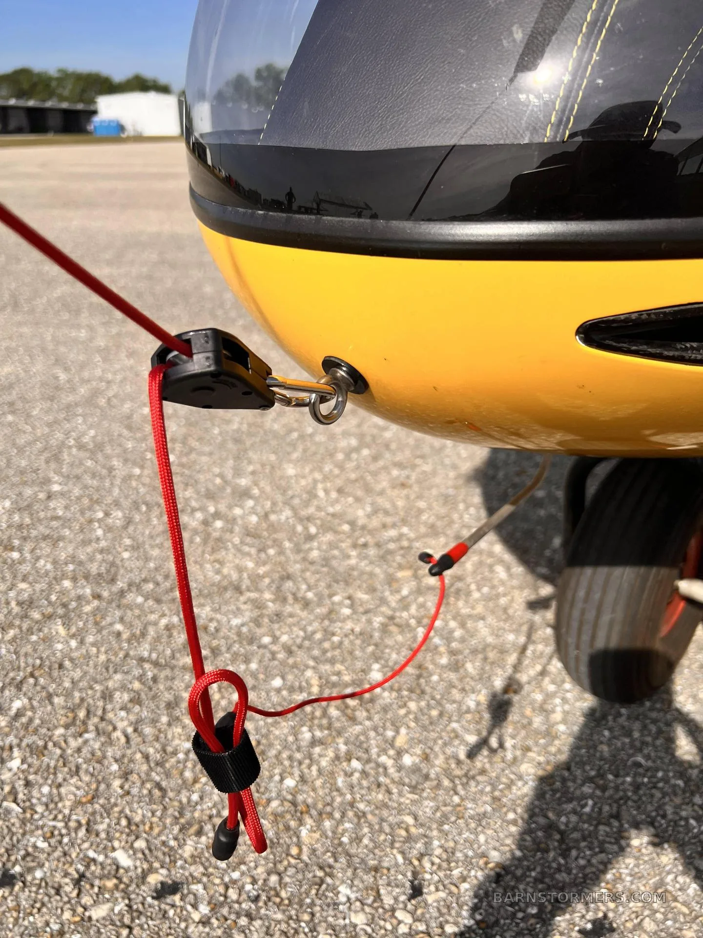 A close-up view of a red tie-down rope secured to the front of a yellow gyrocopter, resting on a gravel surface.