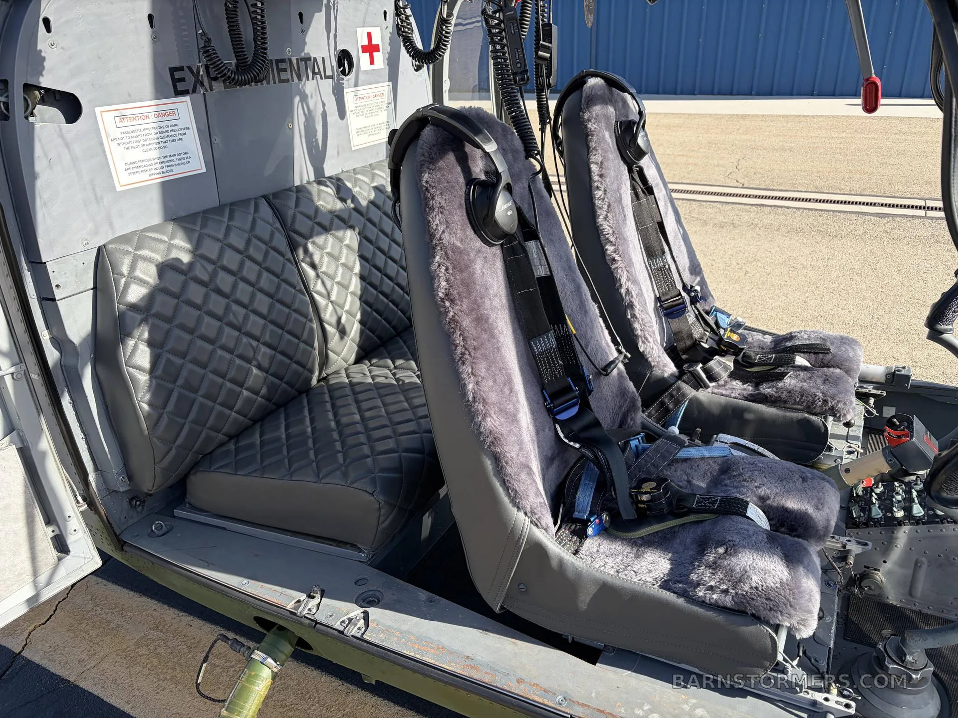 The interior of a helicopter cabin features two front seats with grey sheepskin covers and a quilted grey bench seat in the rear.