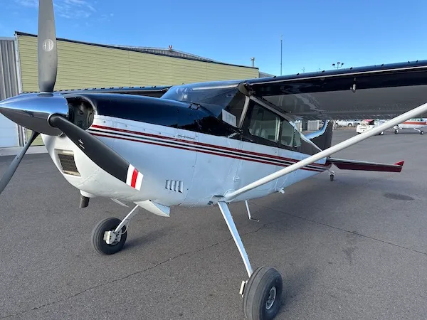 Front-left view of a white and black high-wing propeller aircraft with red stripes, parked on an airfield.