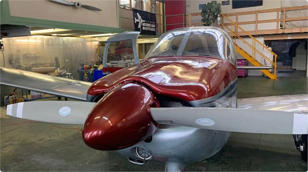 Front view of a red and silver single-engine propeller aircraft with its propeller blades visible, inside a hangar.