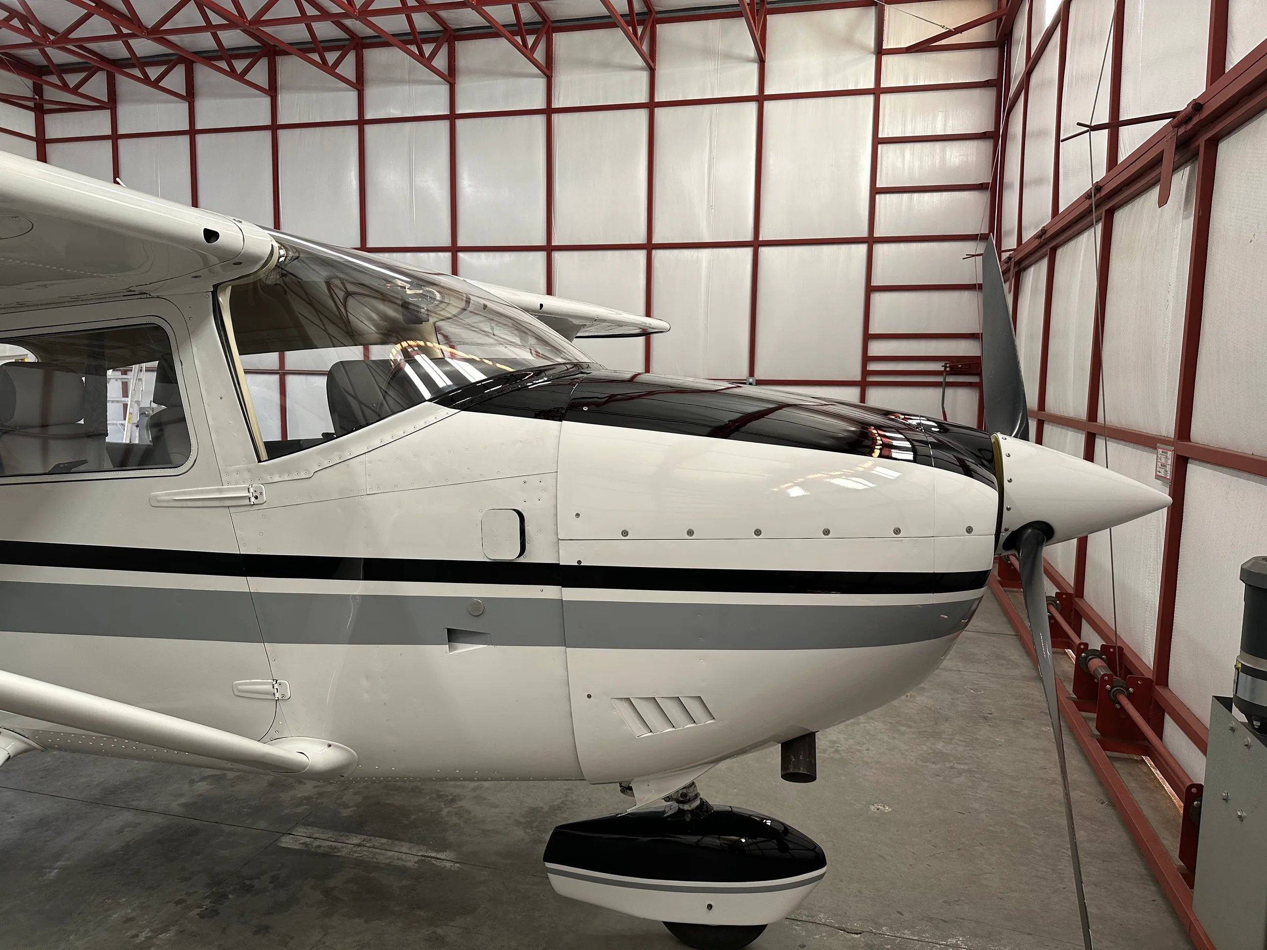 A front-side view of the Cessna 182's nose and propeller, showing its white and grey paint scheme inside a hangar.