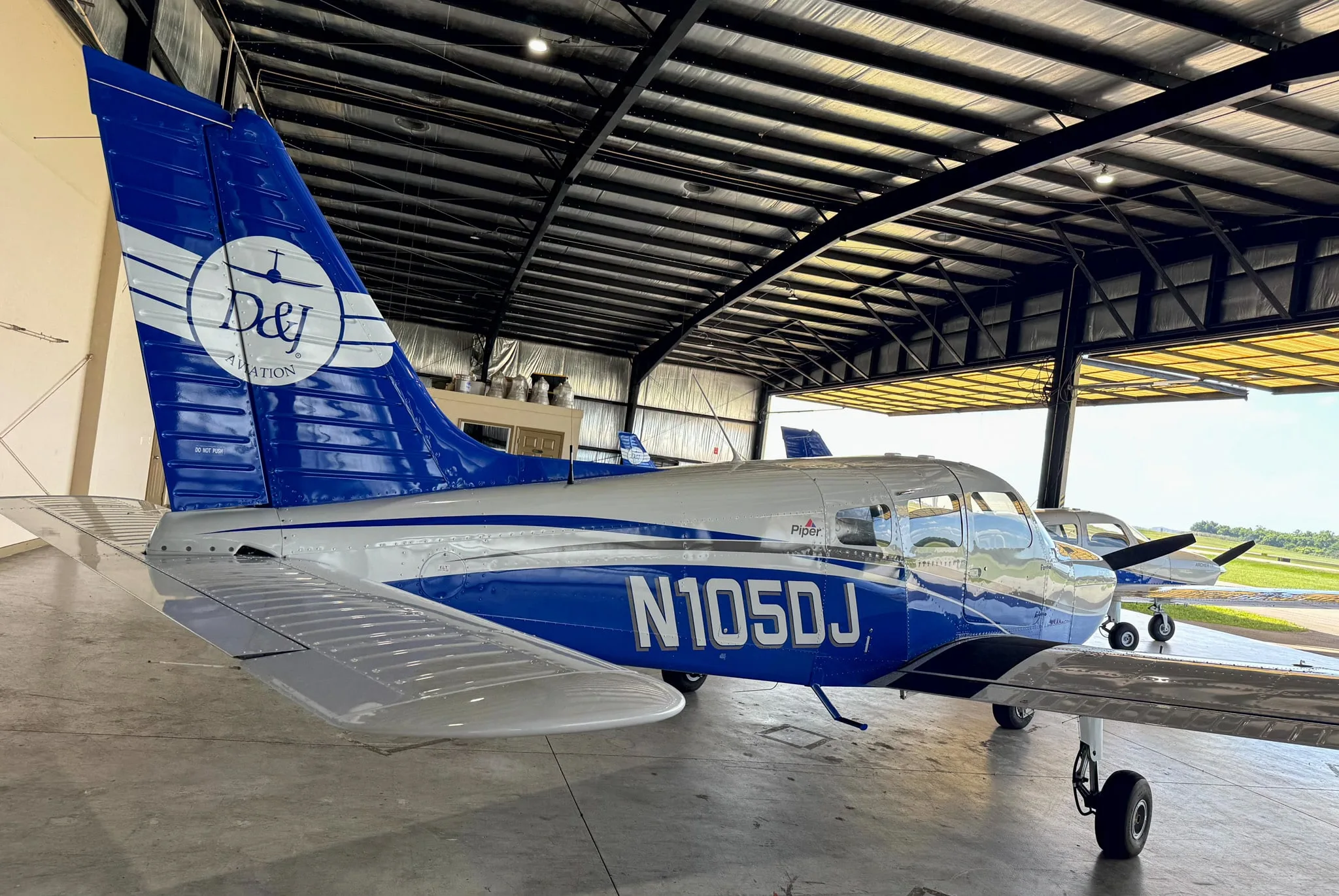 The tail and right side of the white and blue Piper Archer TX, N105DJ, are visible inside a hangar with an open door.