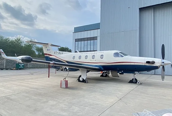 A white and dark blue Pilatus PC-12 turboprop aircraft with a red and gold stripe parked on a tarmac next to a hangar.