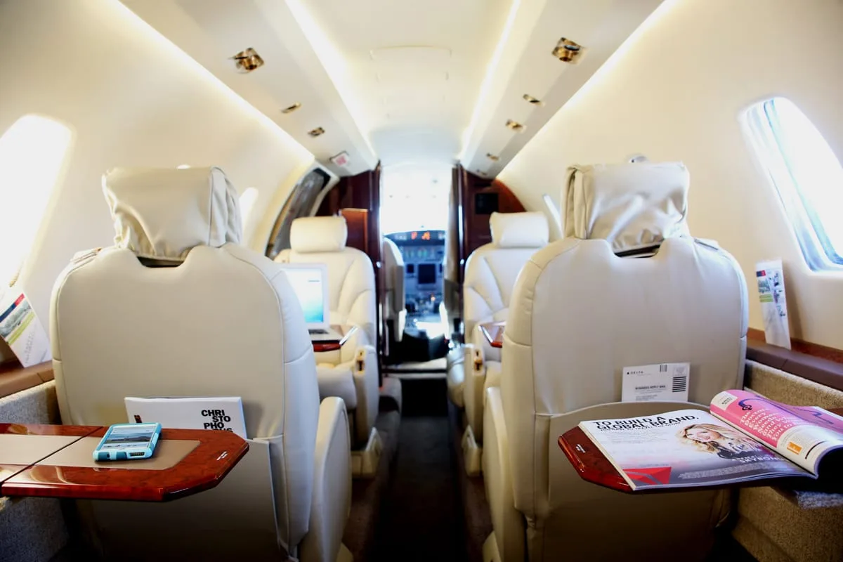 The interior of a private jet cabin features beige leather seats and tables with magazines, looking towards the cockpit.