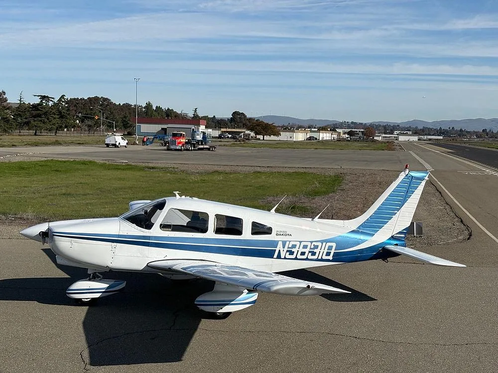 A white Piper Dakota aircraft with blue stripes, registration N3831Q, parked on an airfield with a runway in the background.