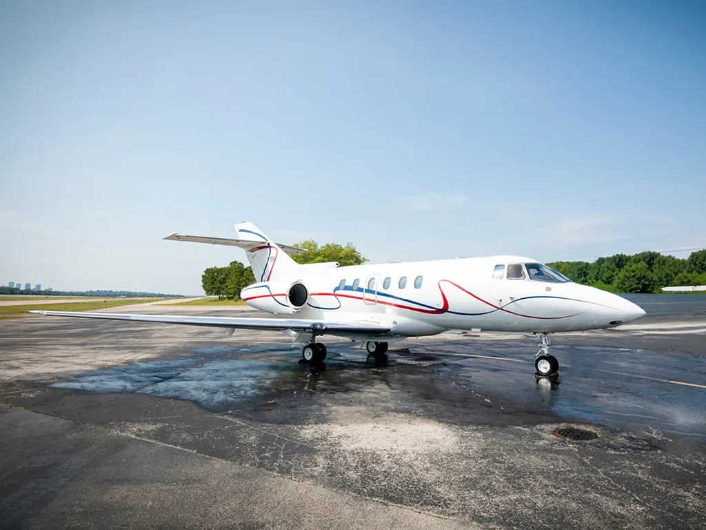 A white business jet with red and blue stripes is parked on a wet tarmac under a clear sky.