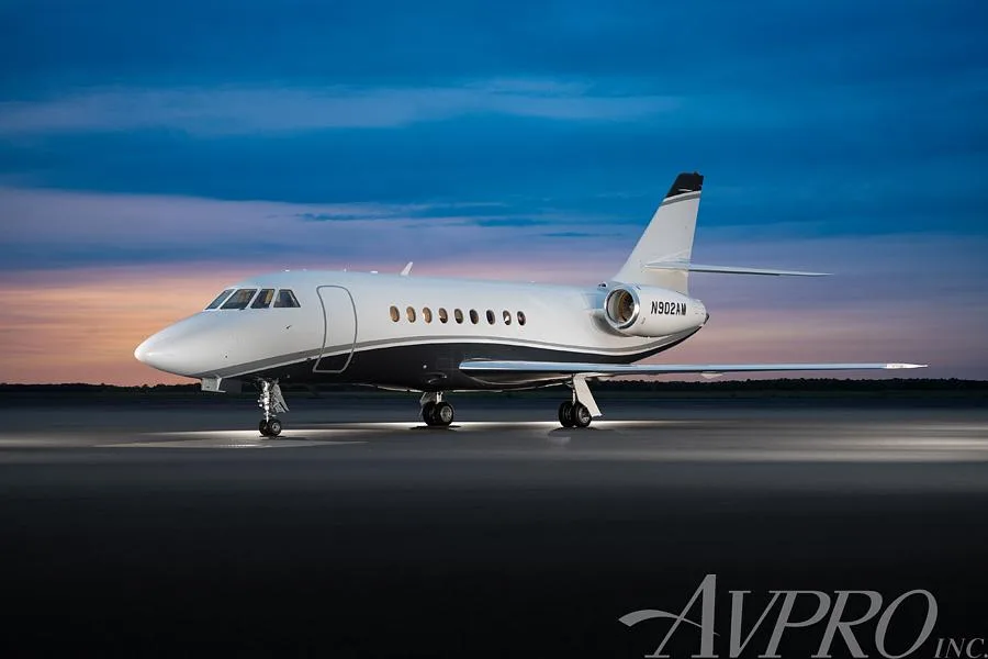 A Dassault Falcon business jet, registration N902AM, parked on an airfield at sunset, showing its white and dark blue paint scheme.