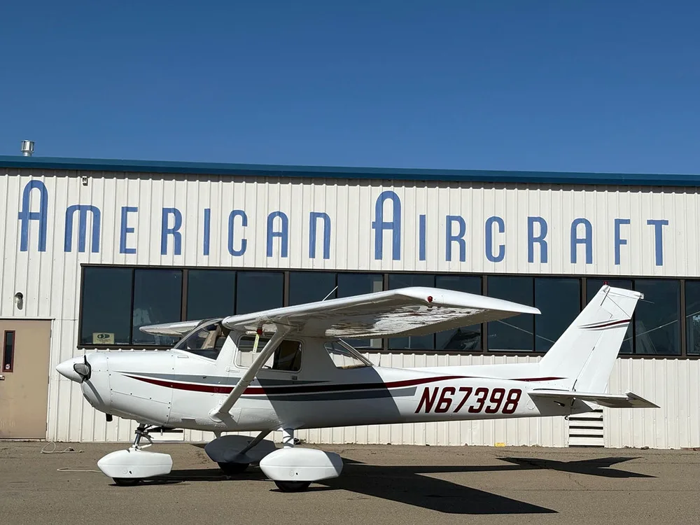 A white Cessna 172 with red and gray stripes, registration N67398, parked on an airfield in front of an "American Aircraft" hangar under a clear blue sky.
