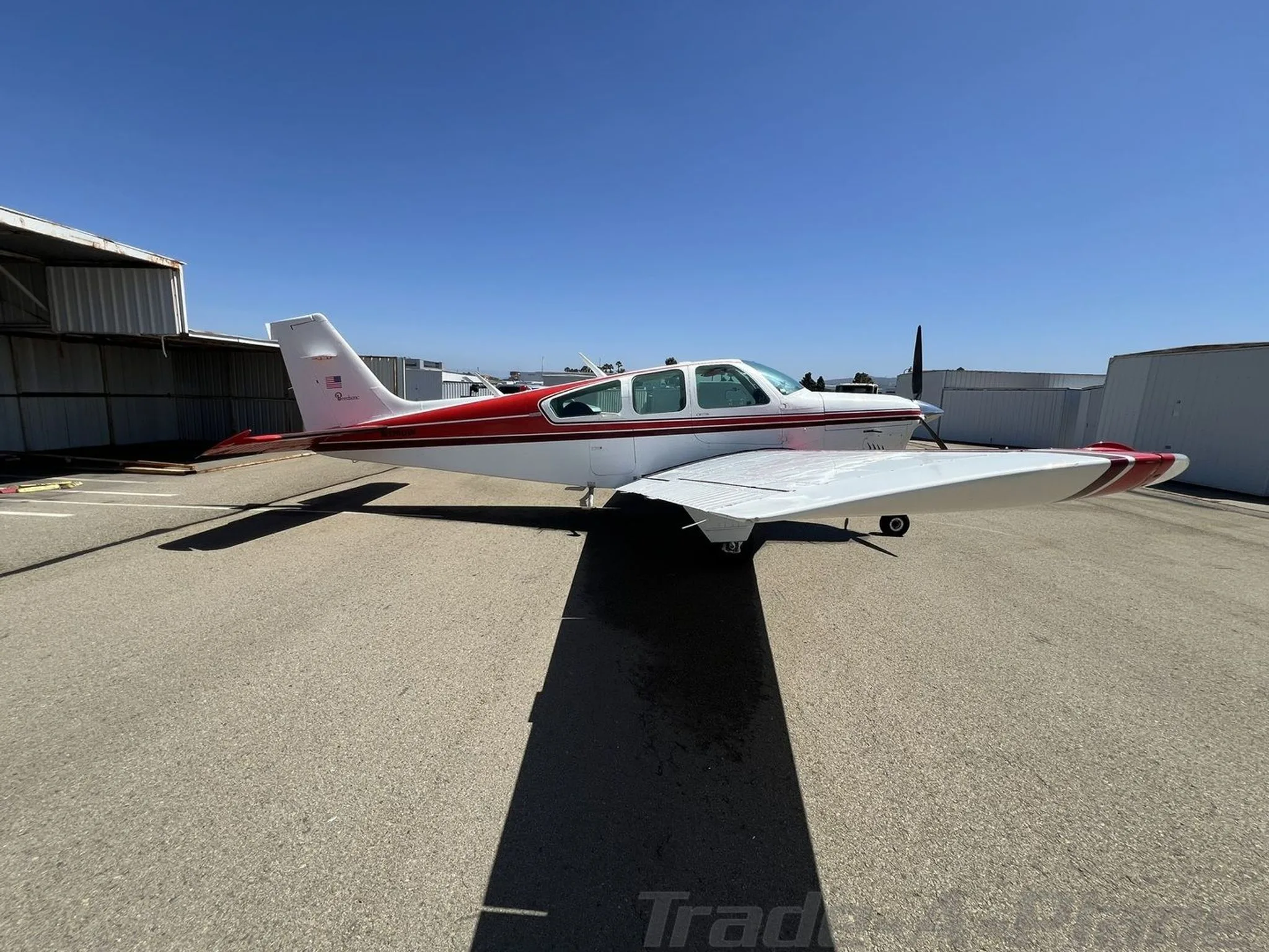 Side view of a white and red Beechcraft Bonanza with a V-tail, parked on an airfield under a clear blue sky.