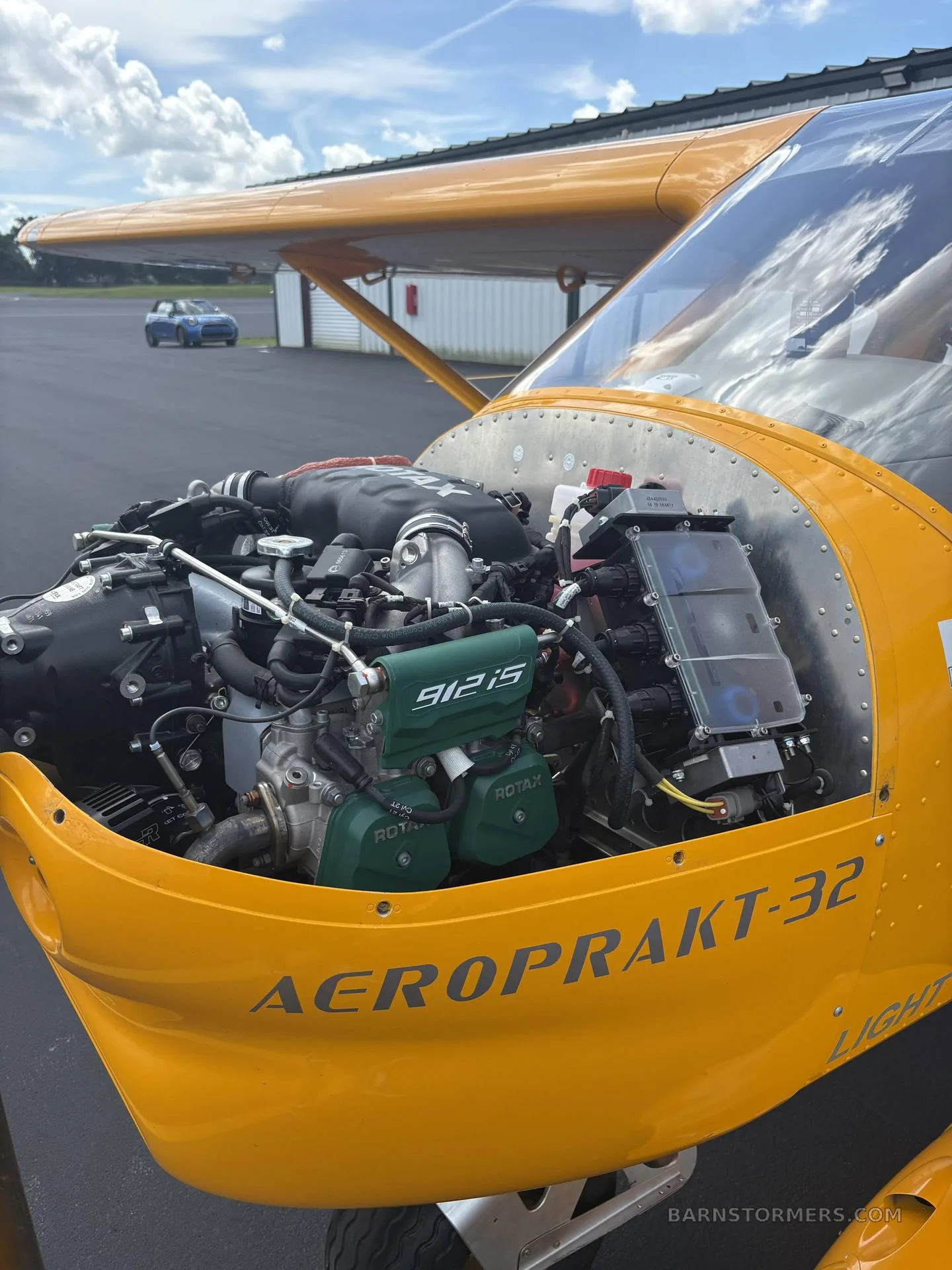 A close-up view of the Rotax 912iS engine exposed in the engine bay of the yellow Aeroprakt A-32 Light Sport aircraft.