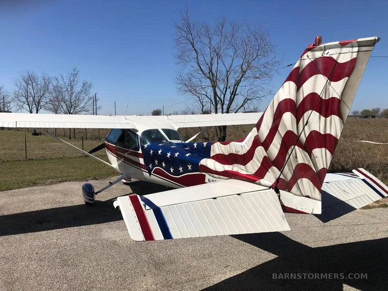 A rear-quarter view of a Cessna 172 with an American flag paint scheme on the fuselage and tail, parked on asphalt.