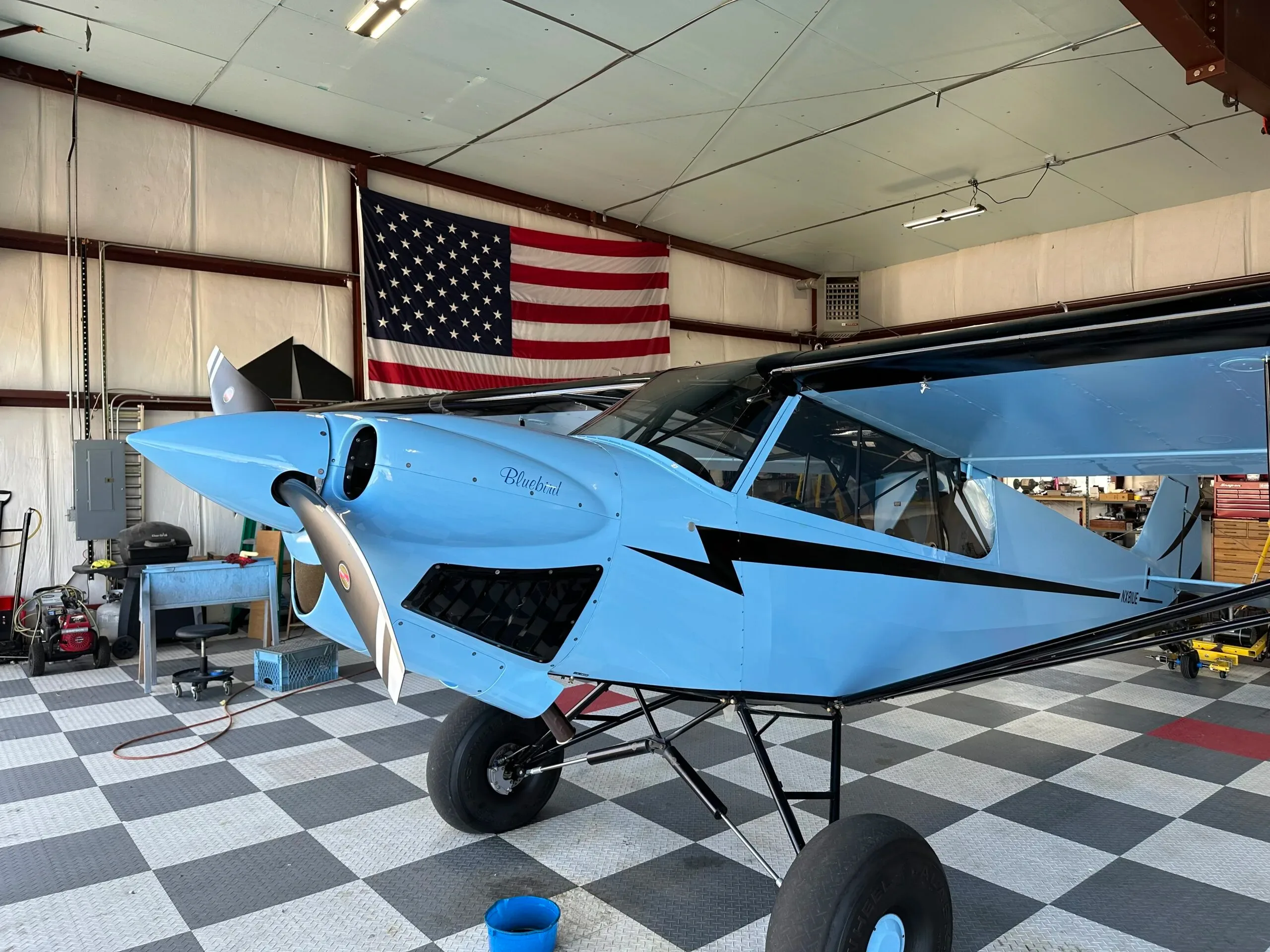 A light blue "Bluebird" aircraft with black accents and large tires, parked in a hangar with an American flag in the background.