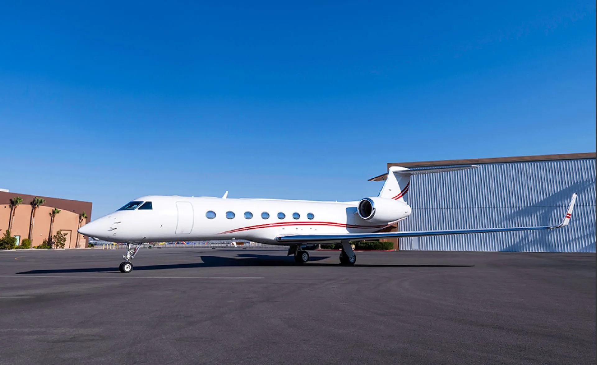 A white Gulfstream private jet with red accent stripes is parked on an airport tarmac in front of a hangar under a clear blue sky.