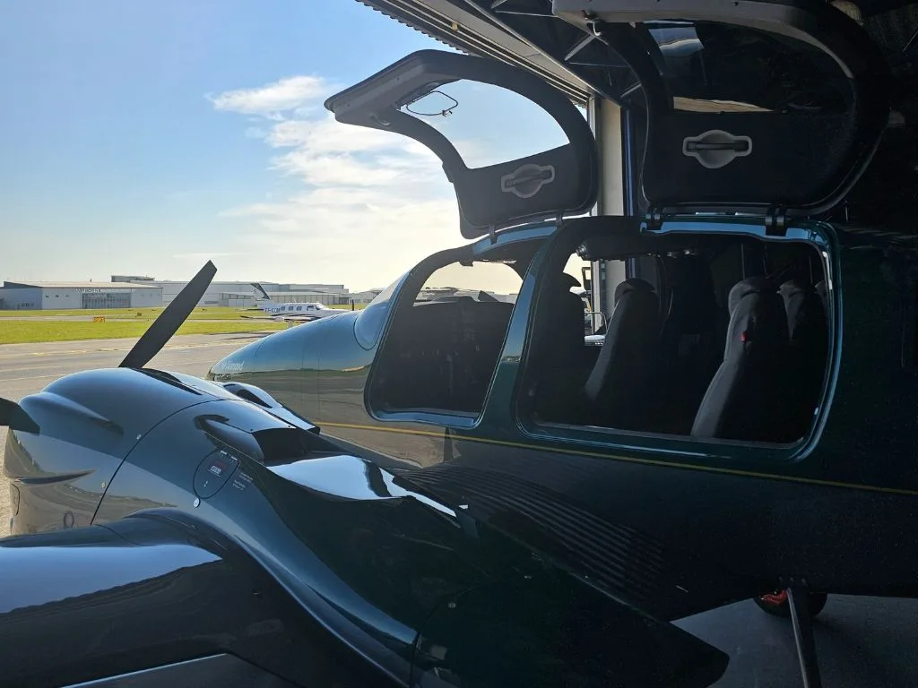 A dark green Diamond DA62 aircraft with its doors open is parked partially inside a hangar, looking out to the airfield.