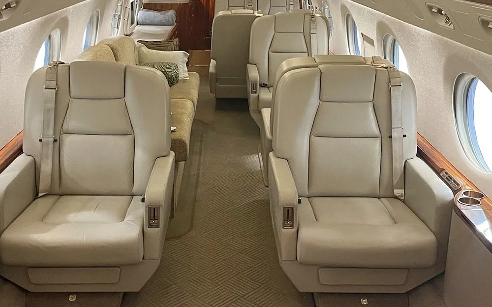 The luxurious cabin interior of a private jet, featuring light beige leather seats arranged in a club configuration and a sofa in the background.