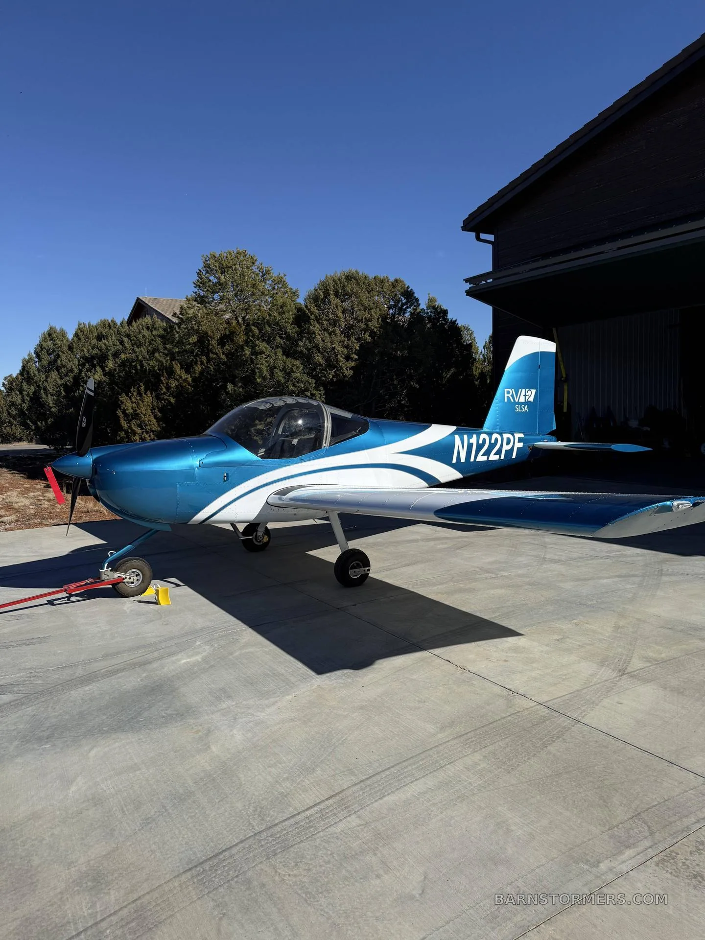 A blue and white RV-12 SLSA aircraft (N122PF) with a red propeller is parked on a concrete pad outside a hangar on a sunny day.