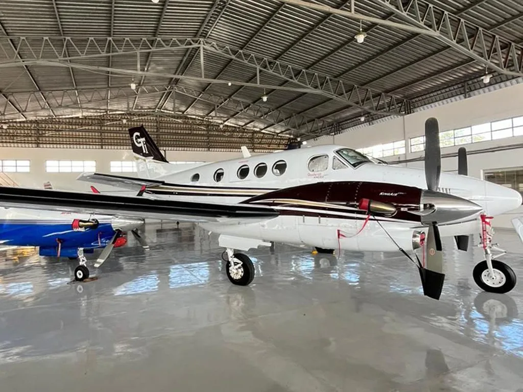 A white and maroon Beechcraft King Air 350 with a 'GT' tail logo, parked in a hangar next to a blue aircraft.