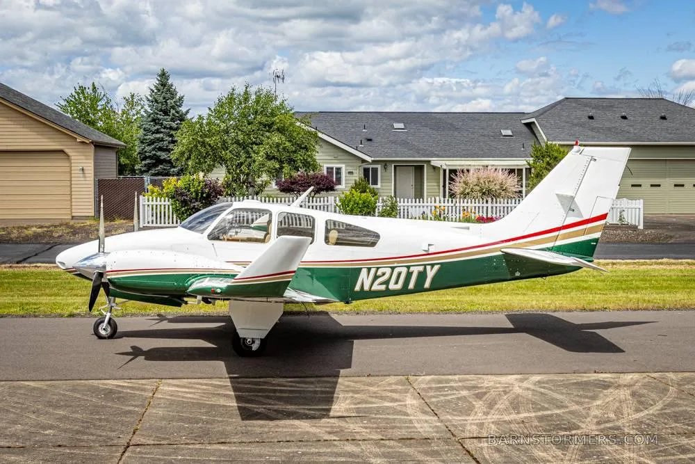 A white and green twin-engine propeller aircraft (N20TY) is parked on a paved surface with houses and trees in the background under a partly cloudy sky.