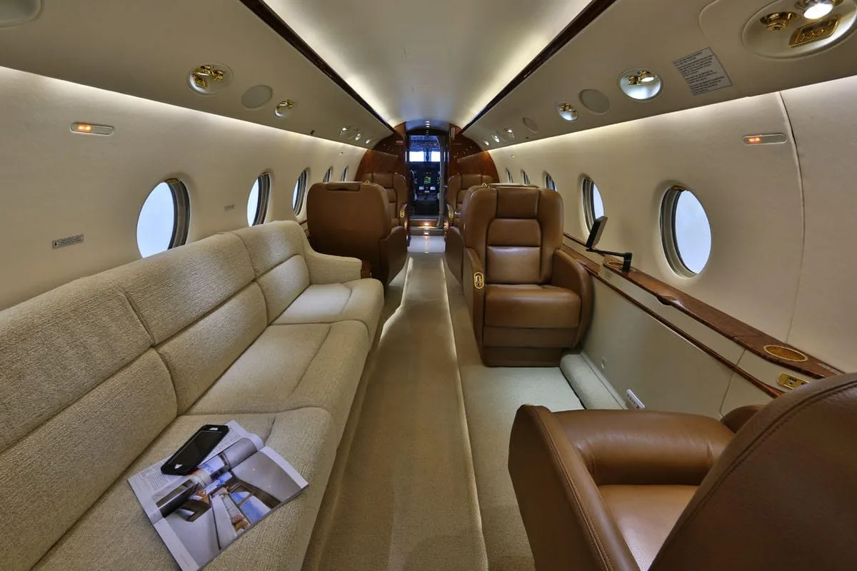 The spacious cabin of a business jet features a long beige sofa, brown leather seats, and oval windows.