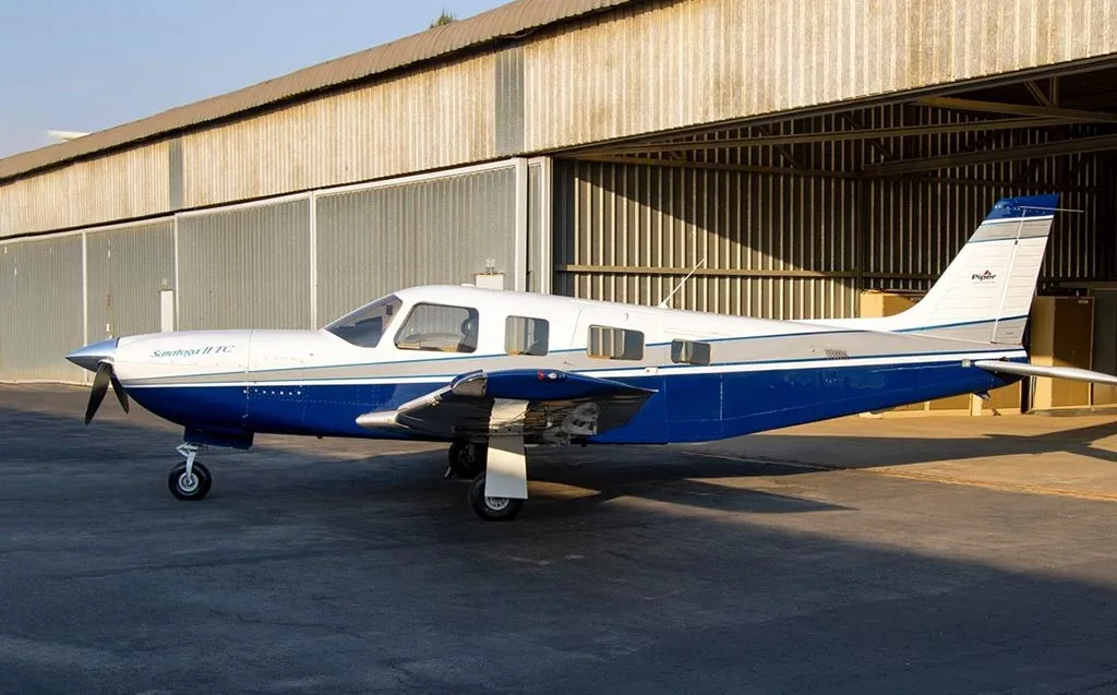 A white and blue Piper Saratoga II TC aircraft parked partially inside an open hangar.