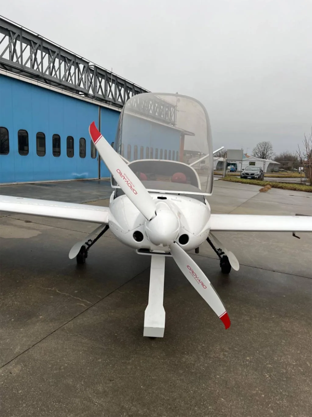 A front view of the white Risen aircraft with a large propeller and open canopy, parked outside a blue hangar.