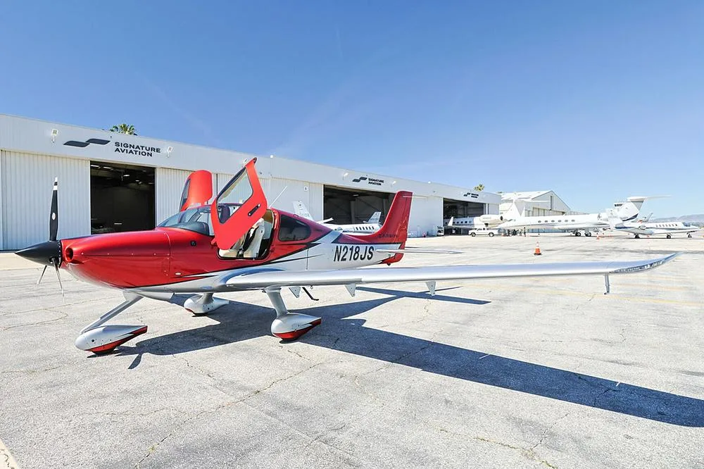 A red and silver Cirrus SR22 aircraft with registration N218JS and open doors, parked on a sunny tarmac.