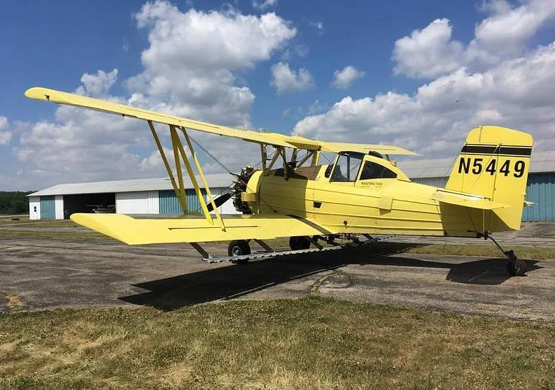 A yellow biplane crop duster, N5449, viewed from the side on an airfield with hangars.
