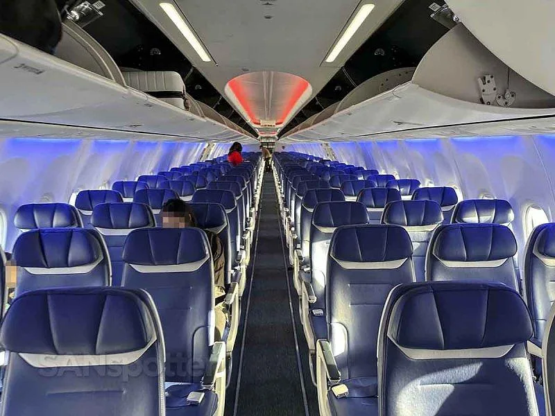 An aircraft cabin with rows of blue seats and overhead bins, illuminated by blue and red ambient lighting.
