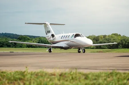 A white Cessna Citation business jet with brown and gold stripes sits on an asphalt runway with trees and hills in the background.