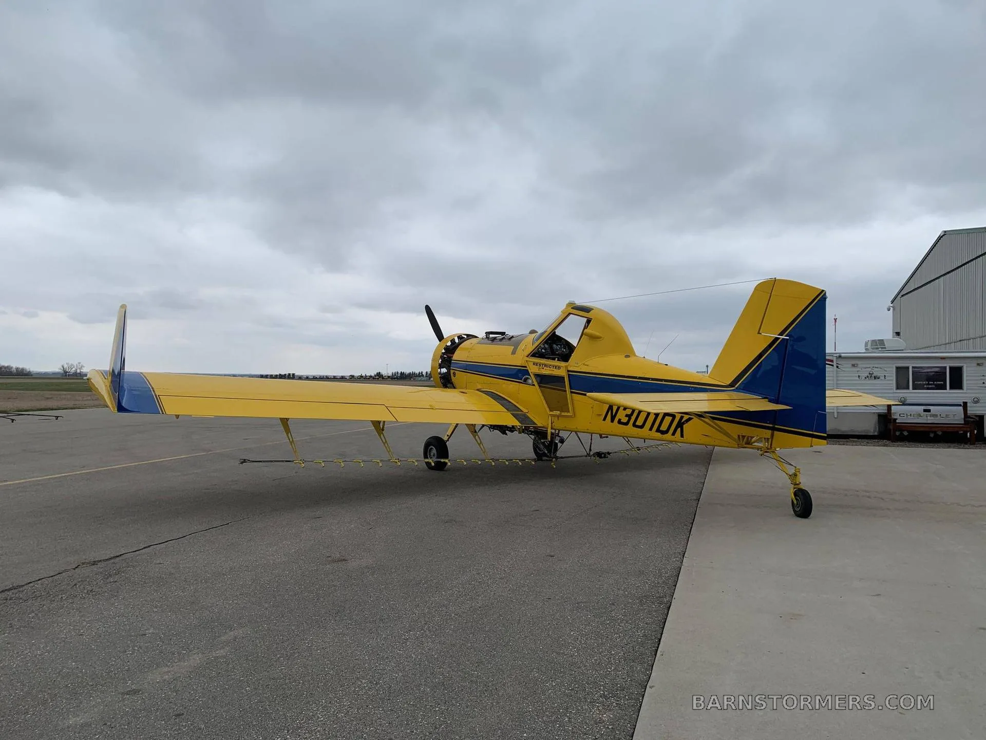 A yellow agricultural aircraft with blue stripes and registration N301DK parked on an airfield under a cloudy sky.