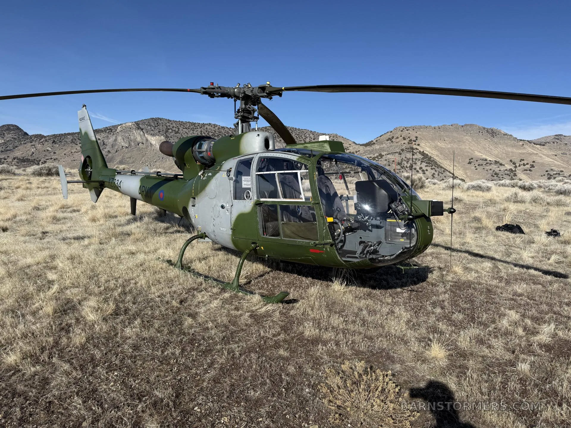 A green and grey military Westland Gazelle helicopter sits on dry, grassy terrain under a clear blue sky.
