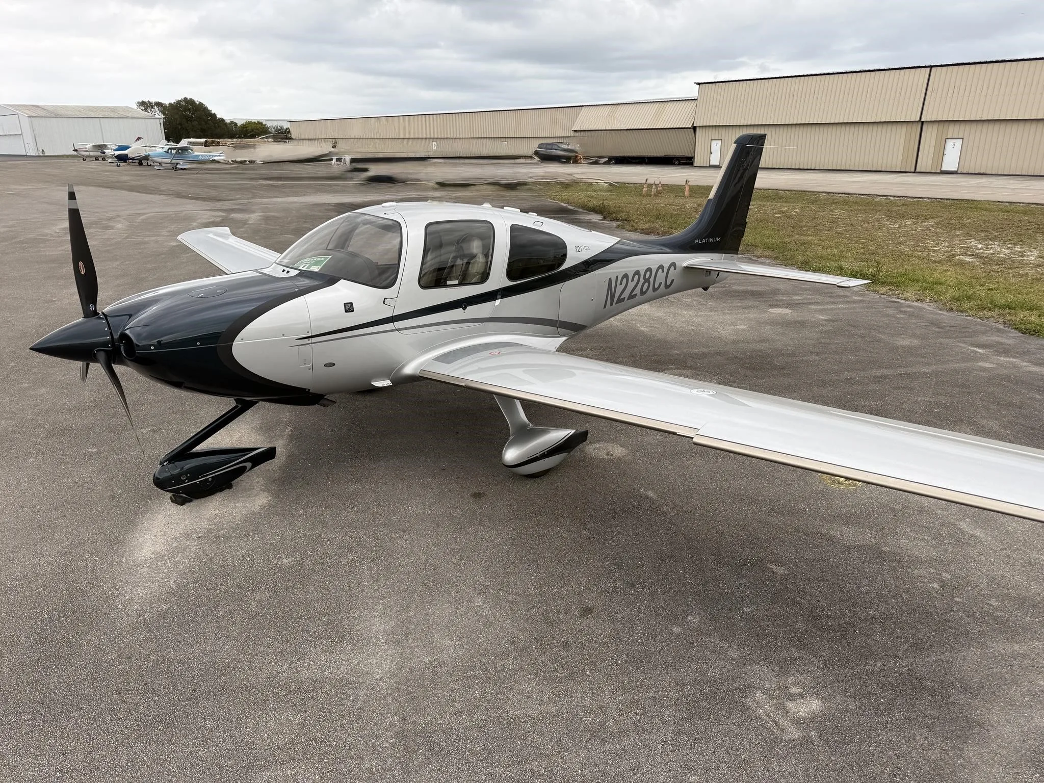 A Cirrus SR22T aircraft, N228CC, with a black and silver paint scheme, parked on an asphalt ramp with hangars in the background.