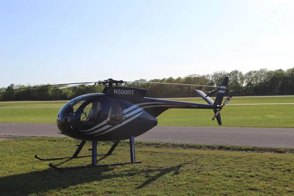 A blue and white Hughes 500 helicopter (N500RT) is parked on a grassy field next to a runway, viewed from the side.