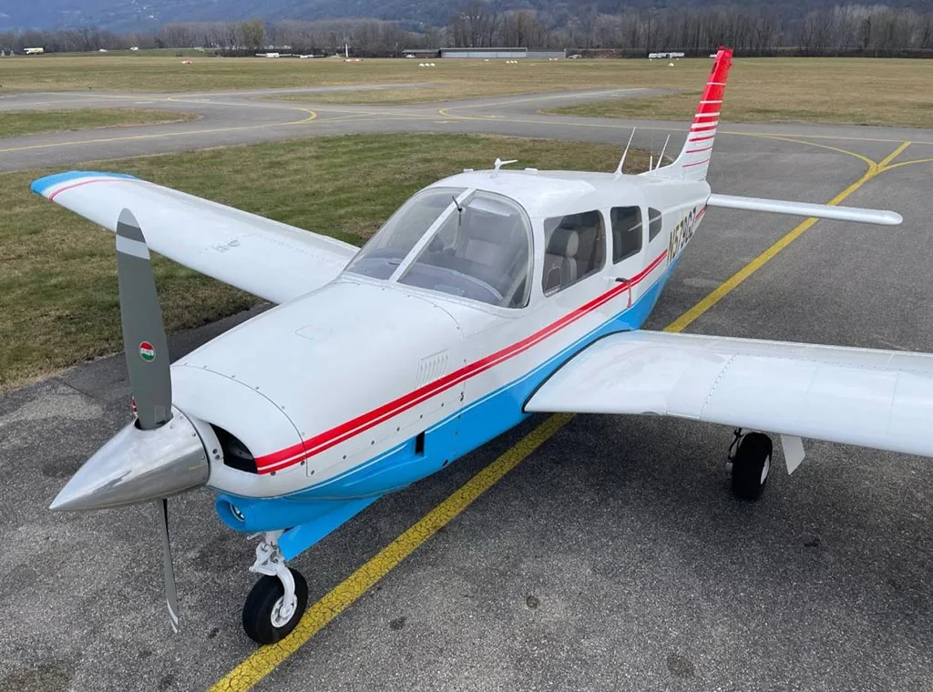 A front-right view of a white and blue Piper PA-28-181 Archer II with red stripes, parked on an airfield.