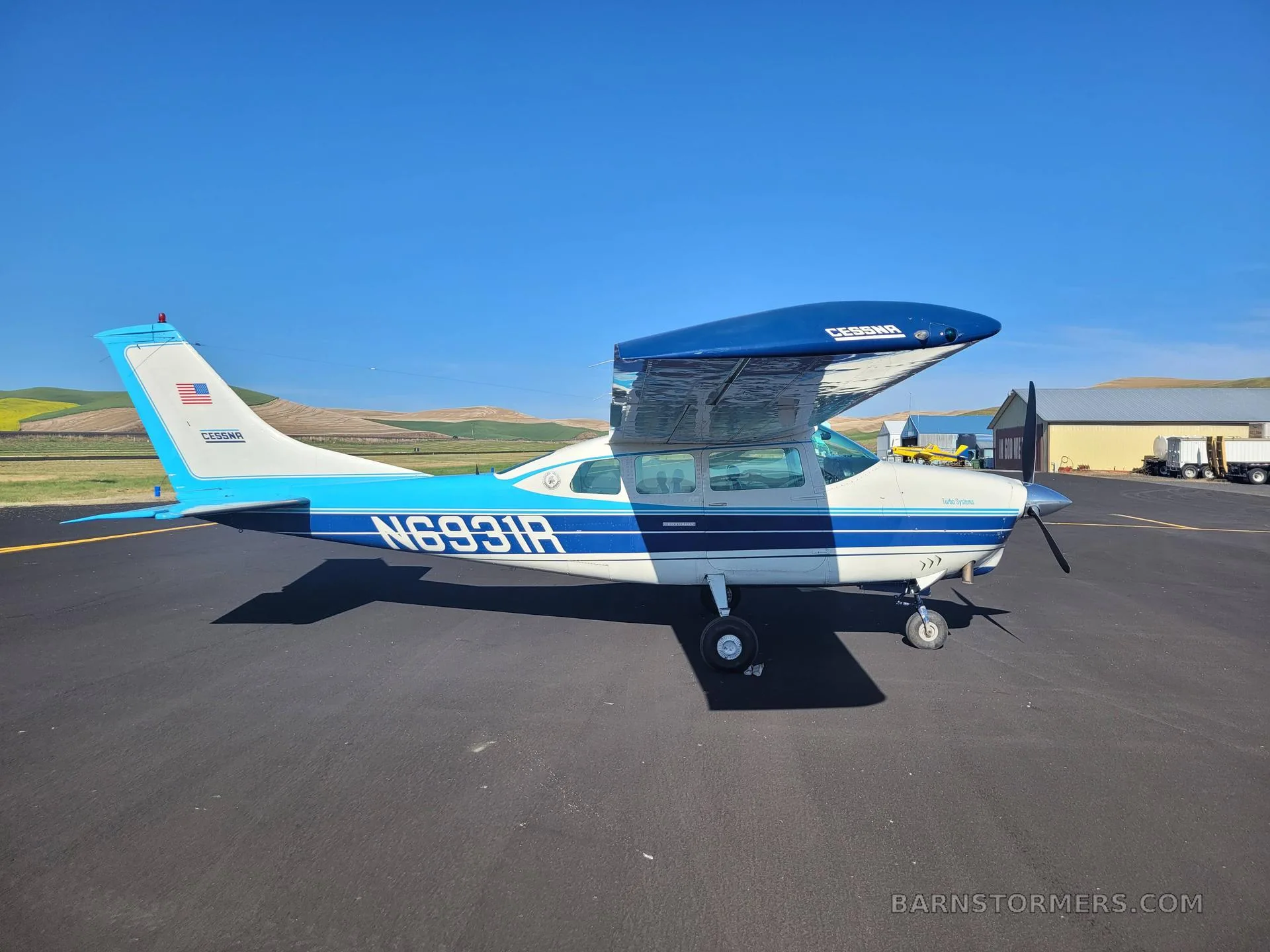 A blue and white Cessna T206H Turbo Stationair (N6931R) parked on an asphalt ramp with rolling hills in the background.