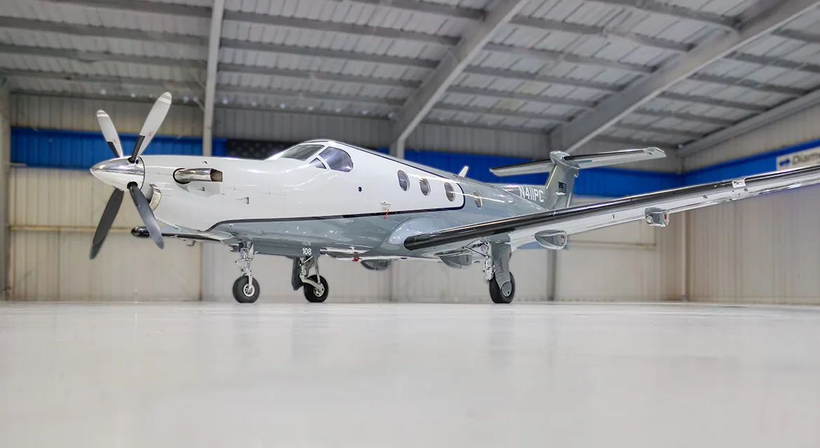 A low-angle view of a white and grey Pilatus PC-12 turboprop aircraft, tail number N411PC, parked inside a hangar.