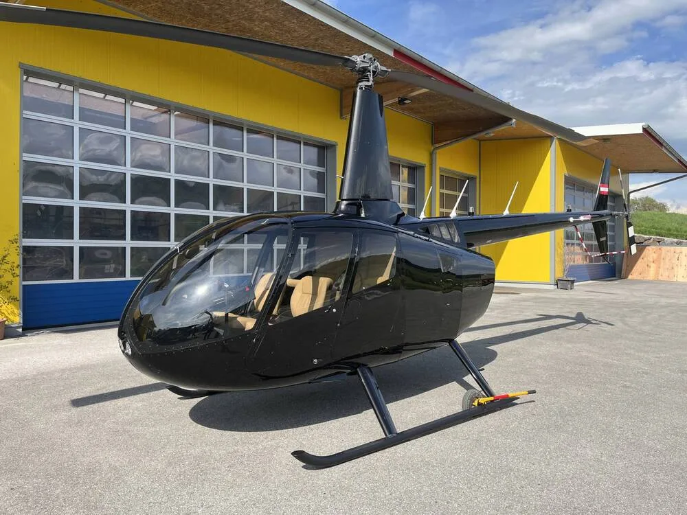 A black Robinson R44 helicopter with tan interior seats is parked on an asphalt apron in front of a yellow hangar.