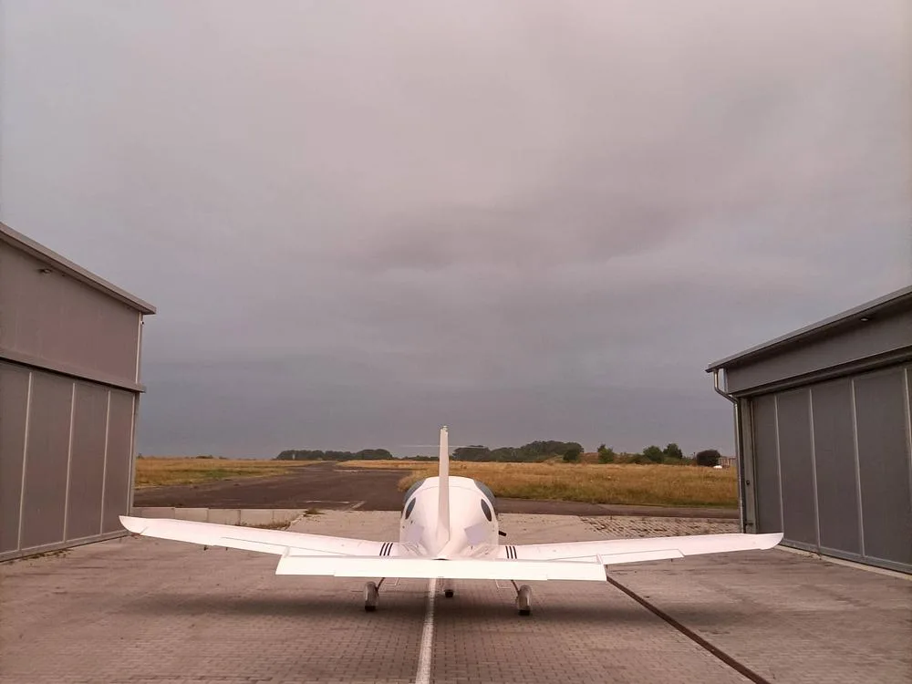 The rear view of a white Bristell aircraft parked on a paved apron, facing a grassy runway under a cloudy sky.