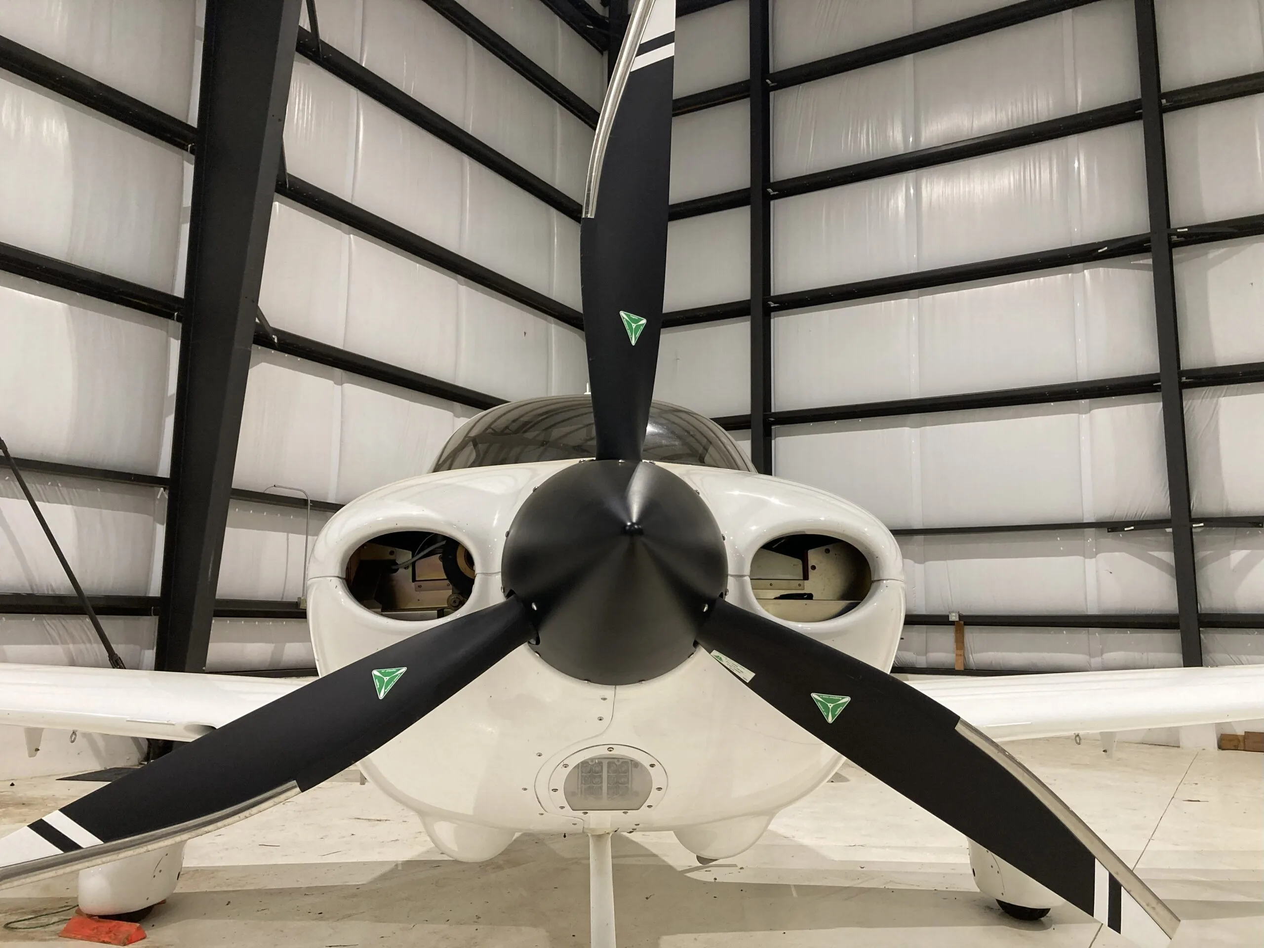 Front view of a white Cirrus SR22 aircraft with a black three-blade propeller, parked in a hangar.