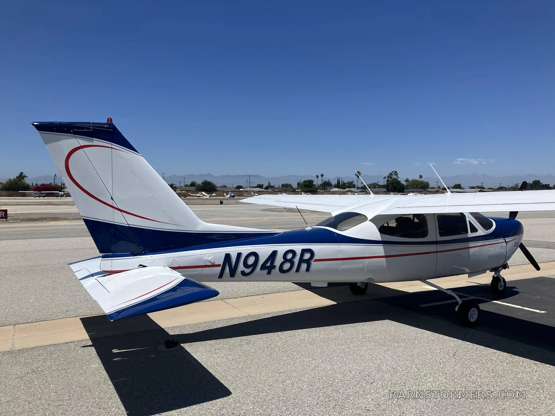A white and blue Cessna 182 Skylane with registration N948R is parked on an asphalt ramp under a clear blue sky, viewed from the rear-right side.