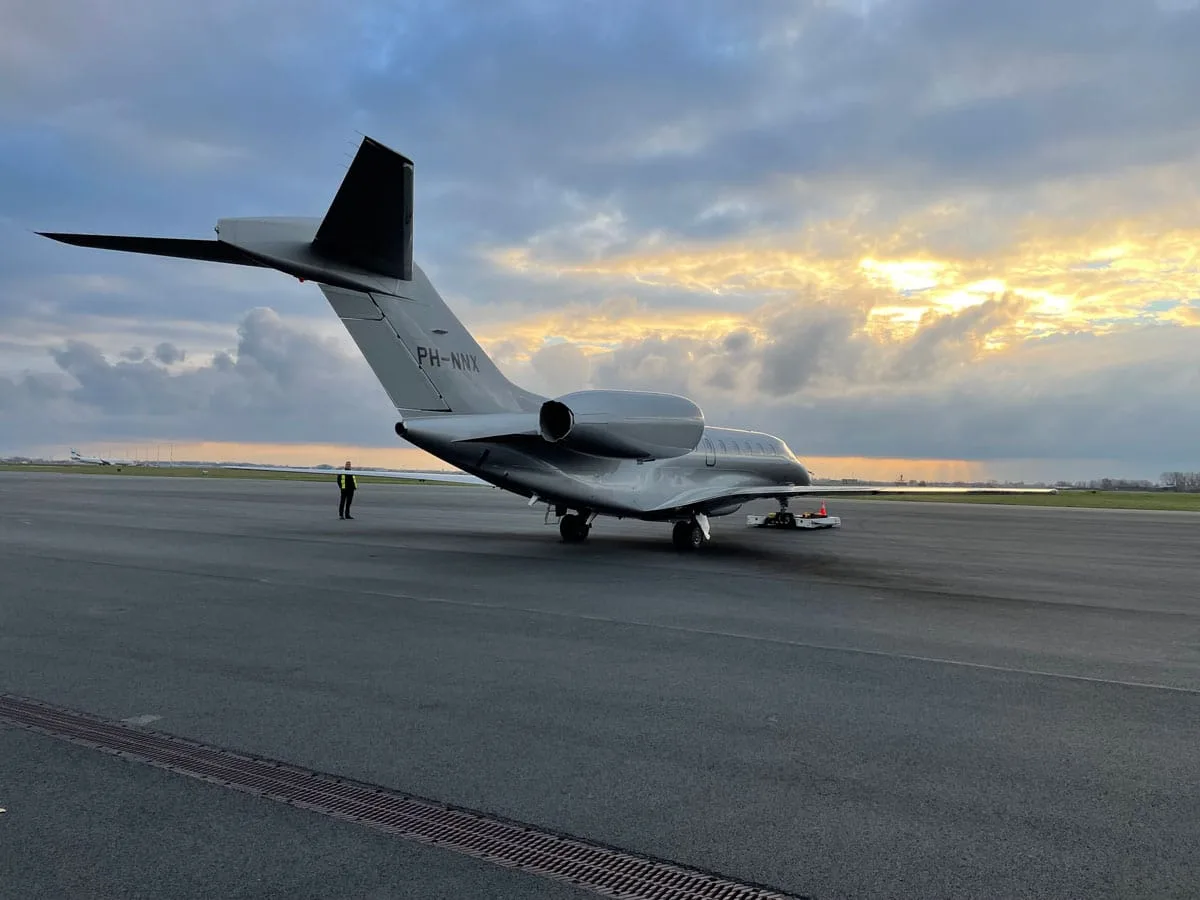 The rear view of a silver private jet (PH-NNX) on an airfield at sunset.