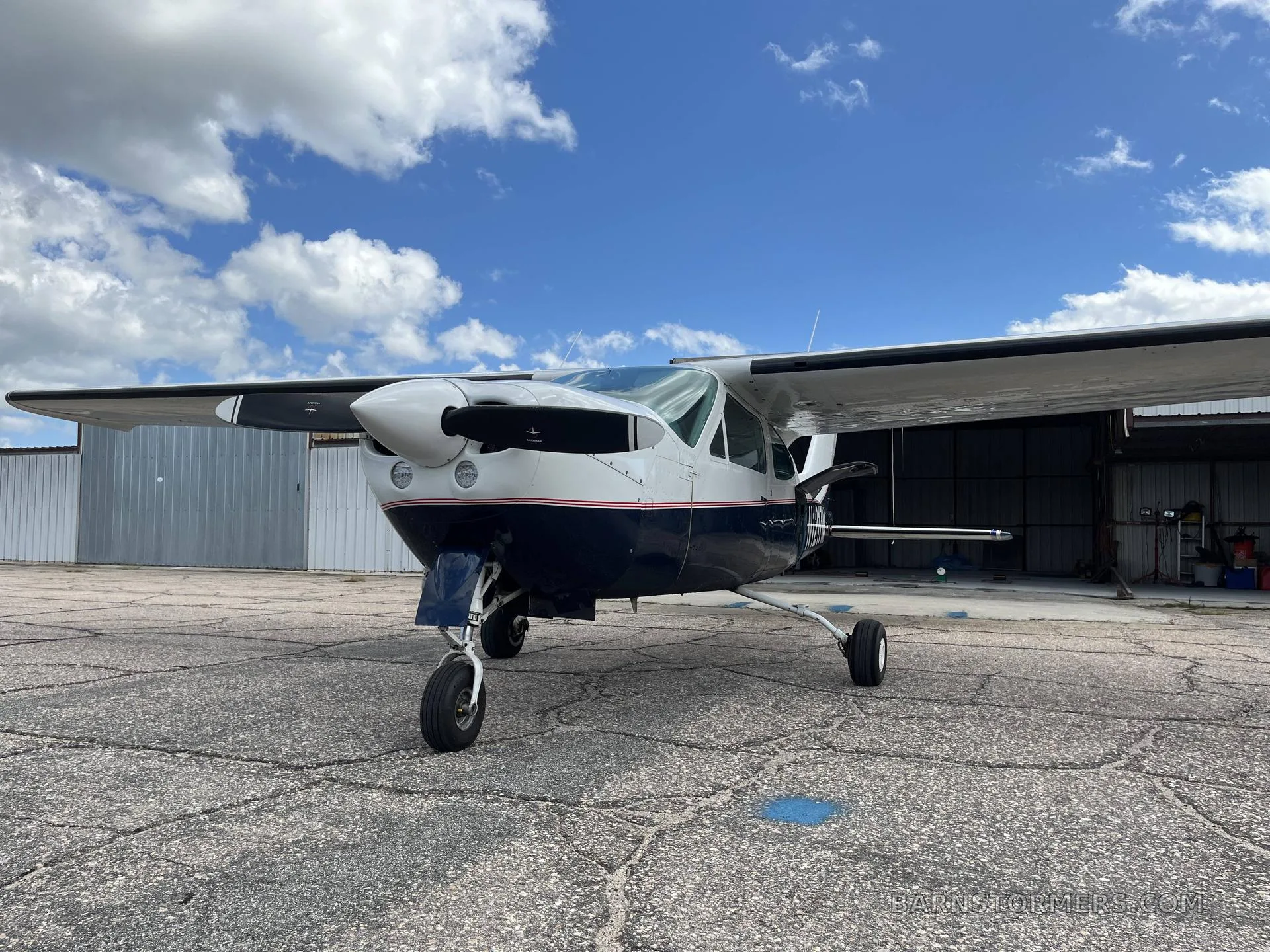 Low-angle front view of the white and dark blue single-engine propeller aircraft N1217M, parked on a concrete apron with hangars in the background.