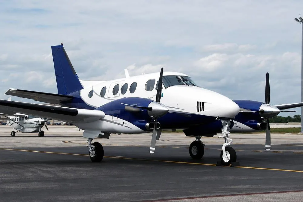 A white and blue twin-engine turboprop aircraft, likely a King Air, is parked on an airport tarmac under a cloudy sky.