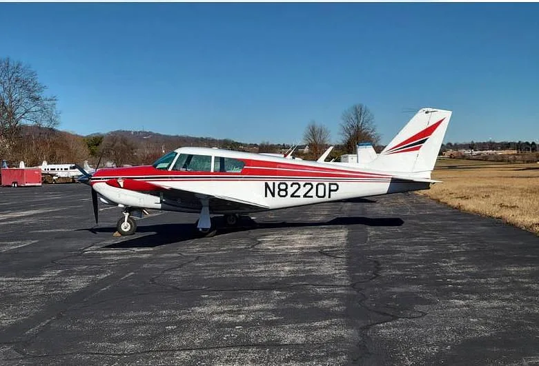 A side view of a red and white Piper Comanche 250 aircraft, tail number N8220P, parked on an airfield.