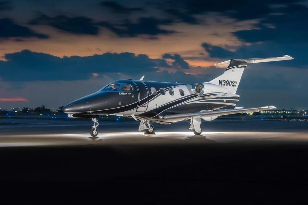 A black and white Embraer Phenom 100 business jet (N390SJ) illuminated on an airport tarmac at sunset.