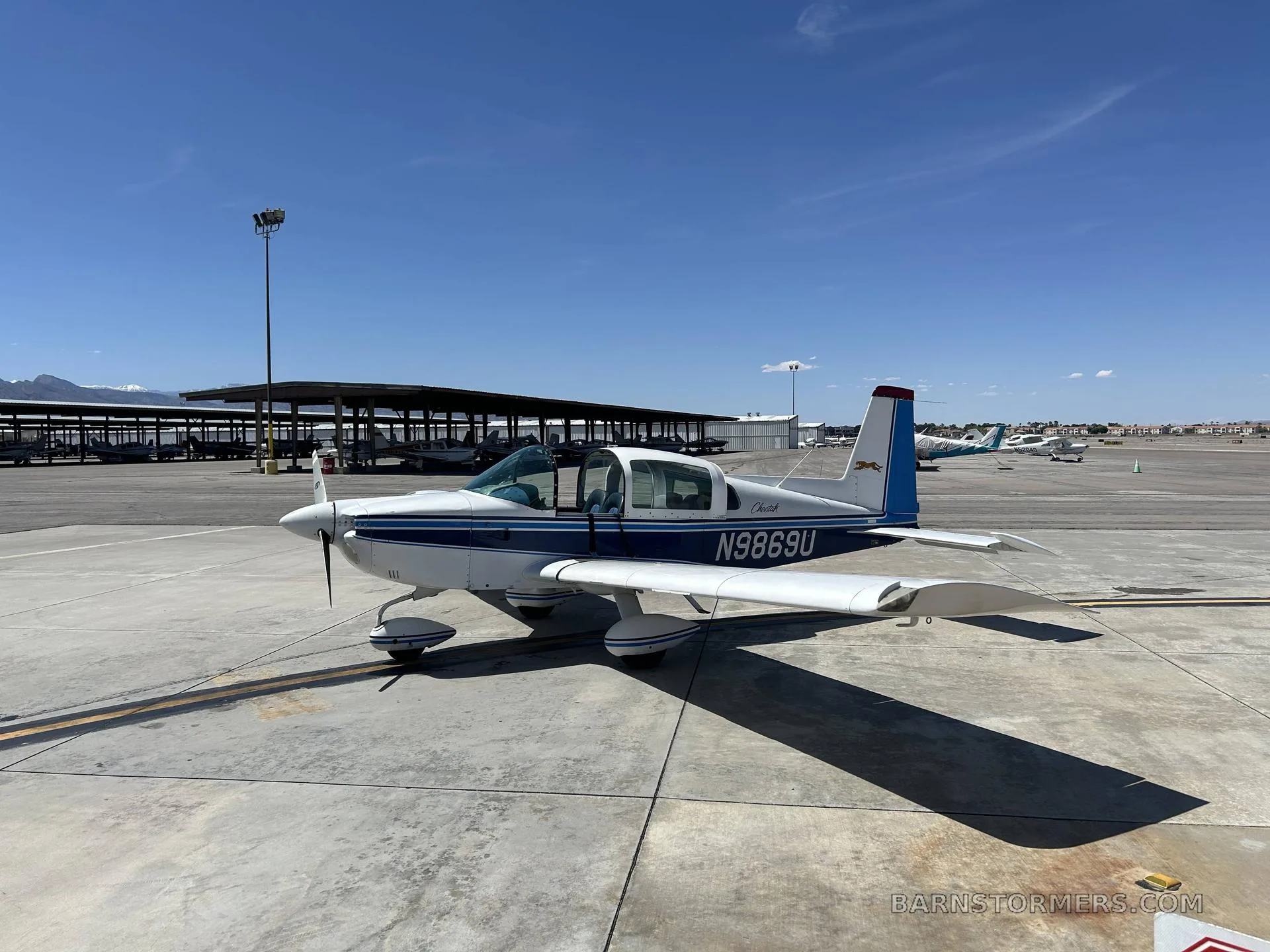 A white and blue Piper Cherokee aircraft (N9869U) is parked on an asphalt tarmac under a clear blue sky, with hangars in the background.