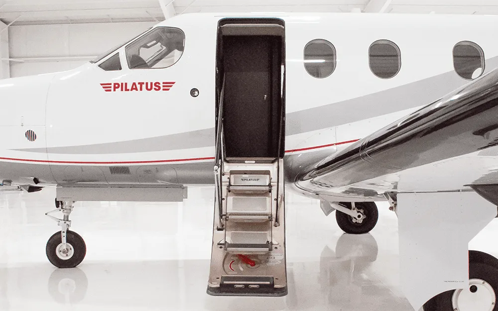 The front section of a white Pilatus PC-12 aircraft with its main passenger door open and integrated stairs extended.